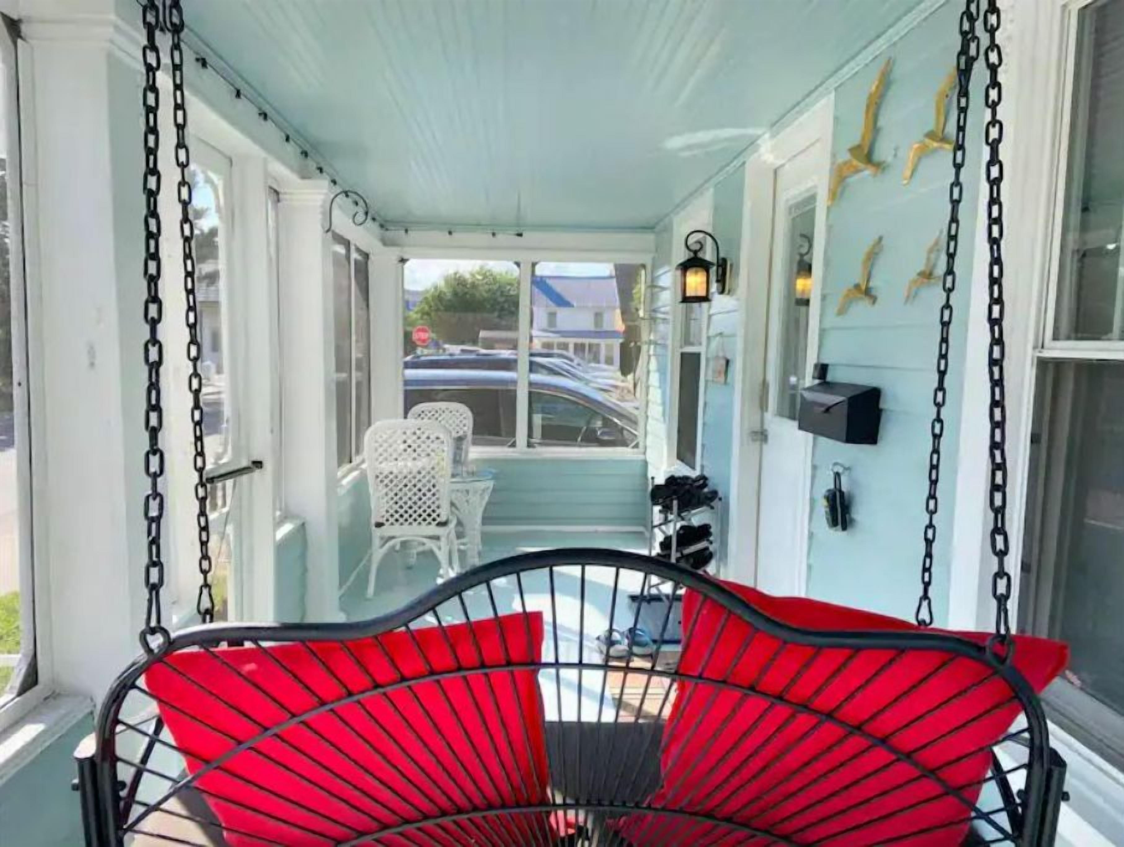 A covered porch with a red swing chair, white chairs, and a view of the street.
