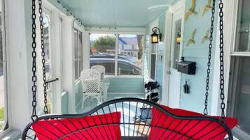 A covered porch with a red swing chair, white chairs, and a view of the street.