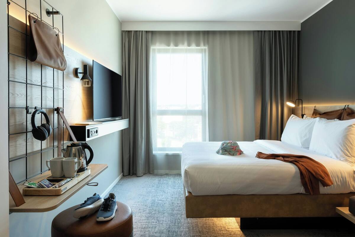 A modern hotel room with a large bed, a TV mounted on the wall, a bedside table with a lamp, and a window with curtains.