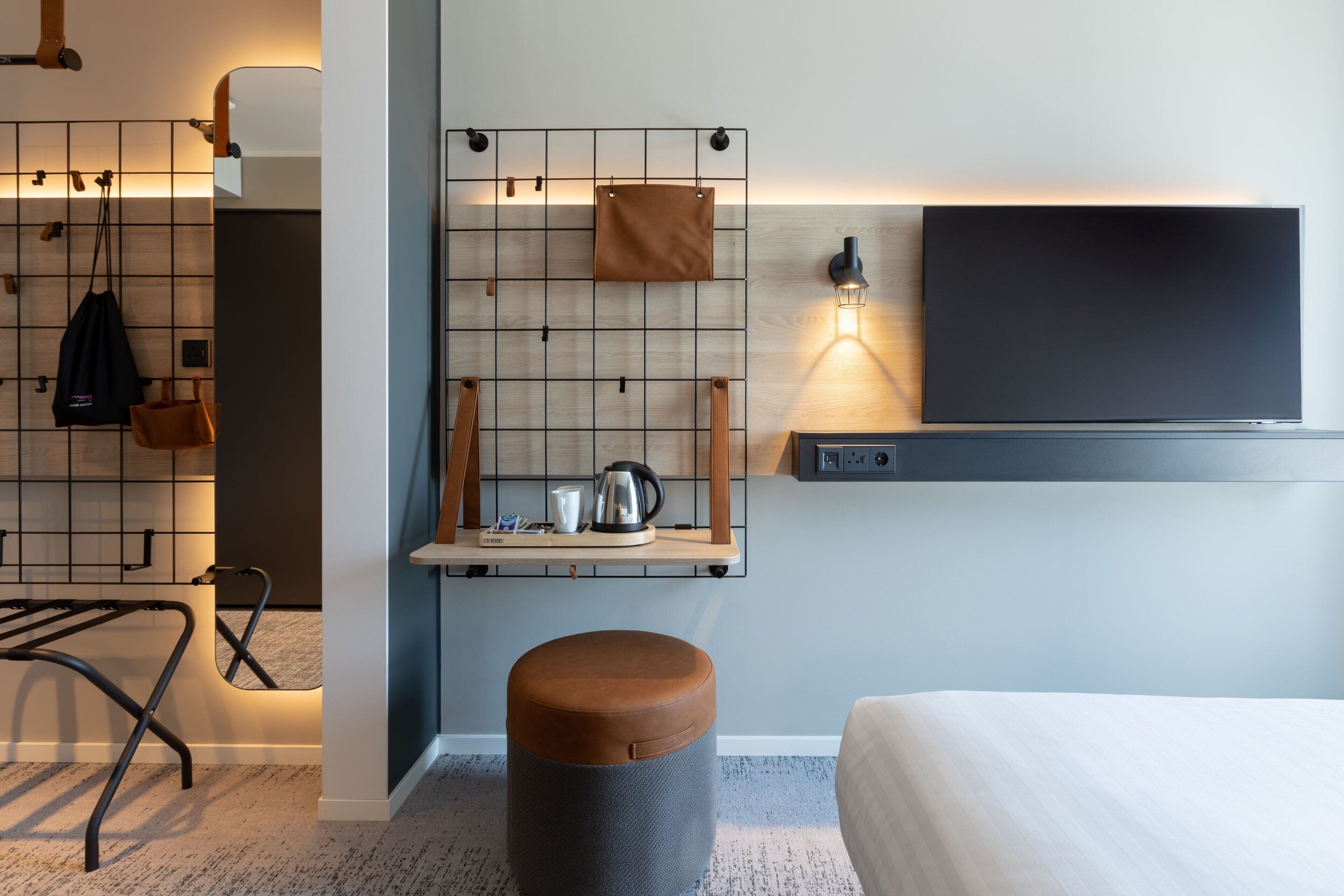 Una habitación de hotel moderna con una cama, una pequeña zona de estar con un taburete, un estante de pared con un hervidor y un televisor.