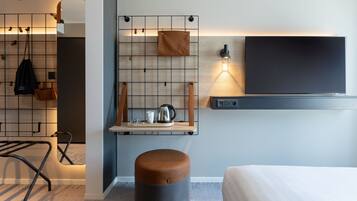 A modern hotel room with a bed, a small seating area with a stool, a wall-mounted shelf with a kettle, and a TV.