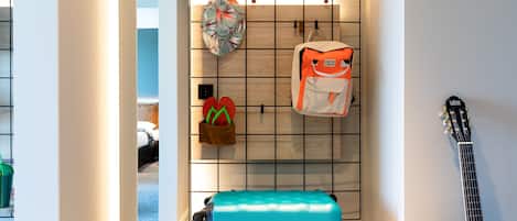 A room with a blue suitcase on a cart, a guitar leaning against the wall, and a wall-mounted rack holding a bag and a hat.