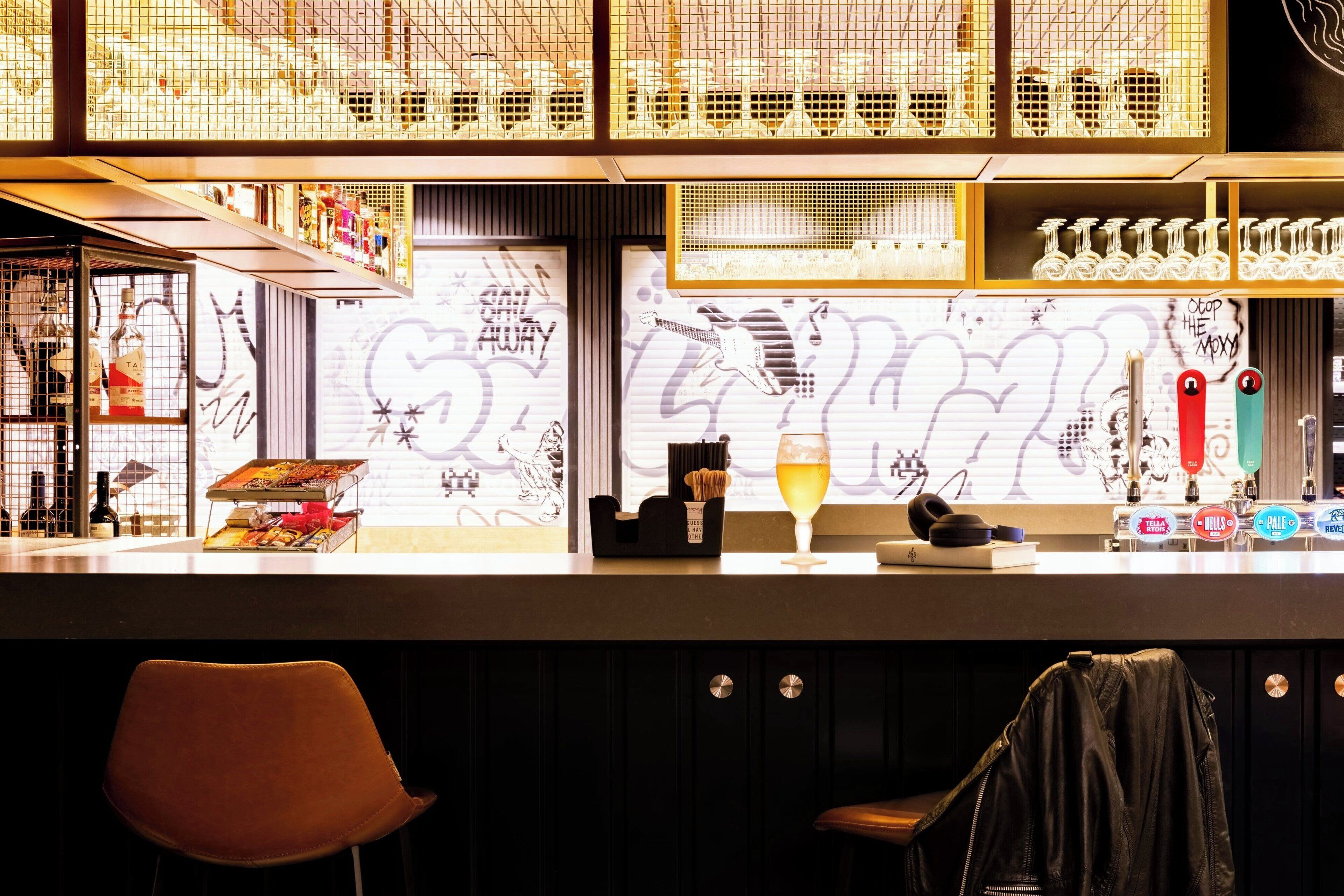A bar area with a counter, shelves with bottles, a glass of beer, and a graffiti wall in the background.