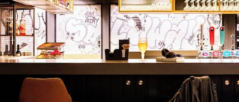 A bar area with a counter, shelves with bottles, a glass of beer, and a graffiti wall in the background.