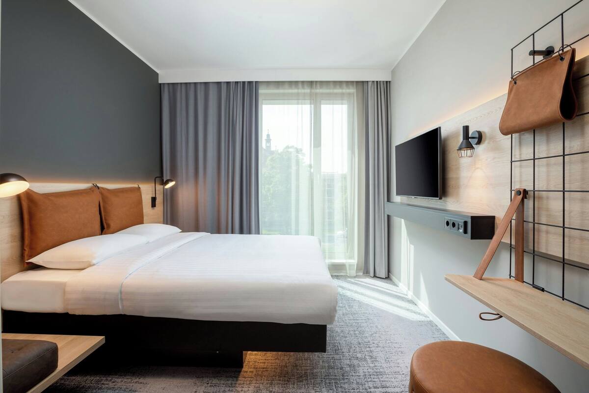 A modern hotel room with a large bed, a TV mounted on the wall, a desk, and a window with curtains.