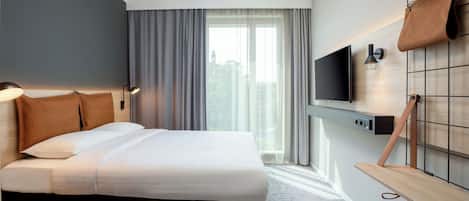 A modern hotel room with a large bed, a TV mounted on the wall, a desk, and a window with curtains.