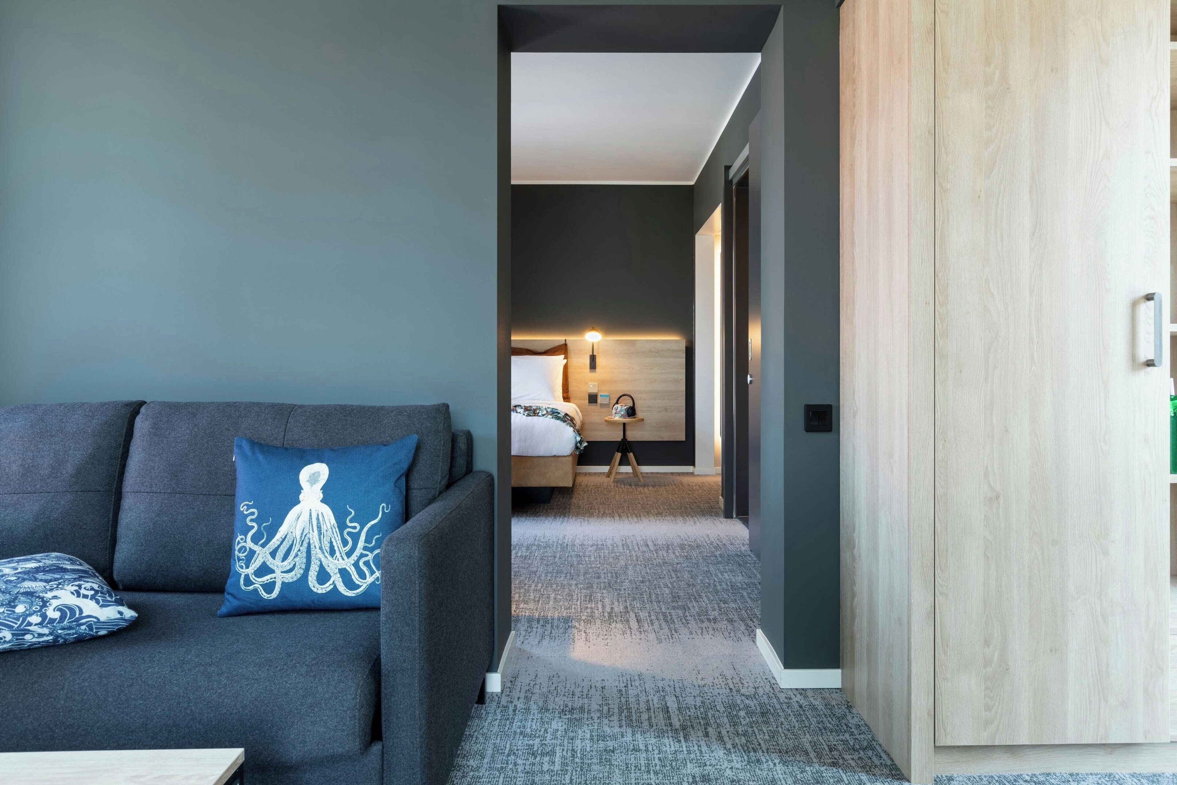 Una habitació d'hotel moderna amb un sofà gris, un coixí blau amb forma d'pop i un armari de fusta, i un llit ben fet.