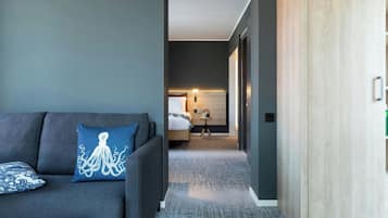 A modern hotel room with a grey sofa, a blue octopus pillow, a wooden wardrobe, and a neatly made bed.