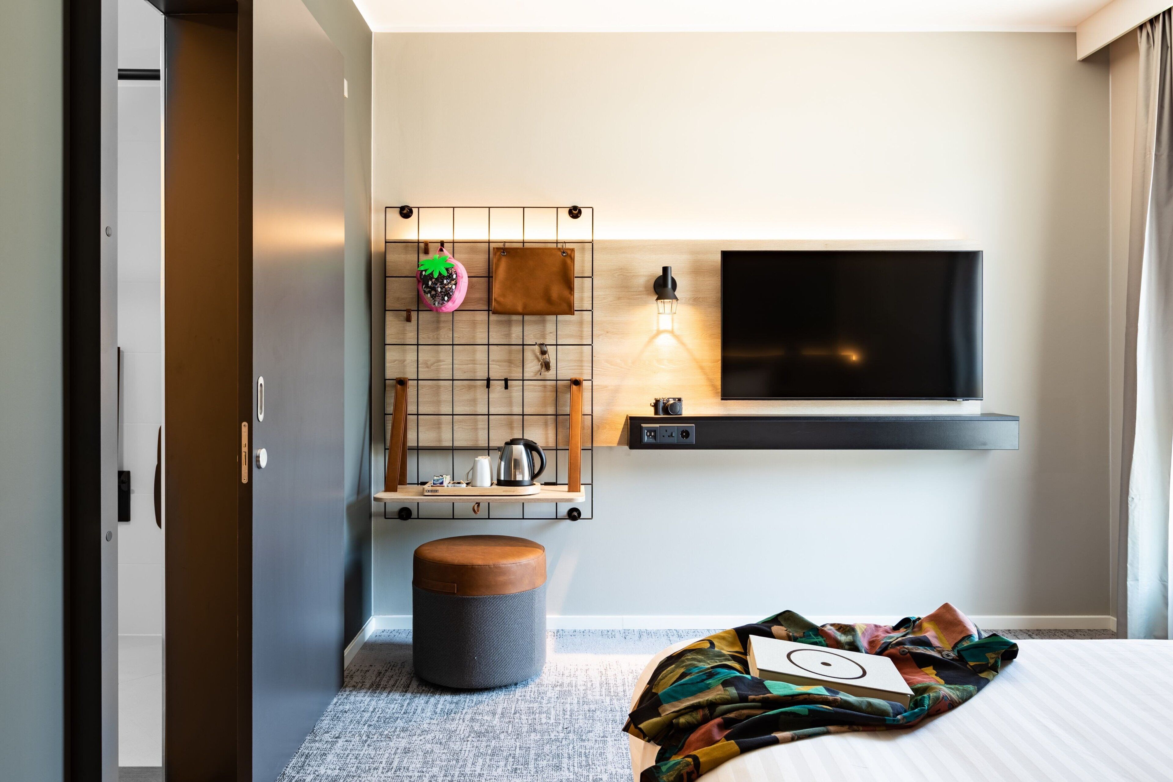 A modern hotel room with a large bed, a TV mounted on the wall, a small table with a kettle, and a unique wall-mounted shelf with decorative items.