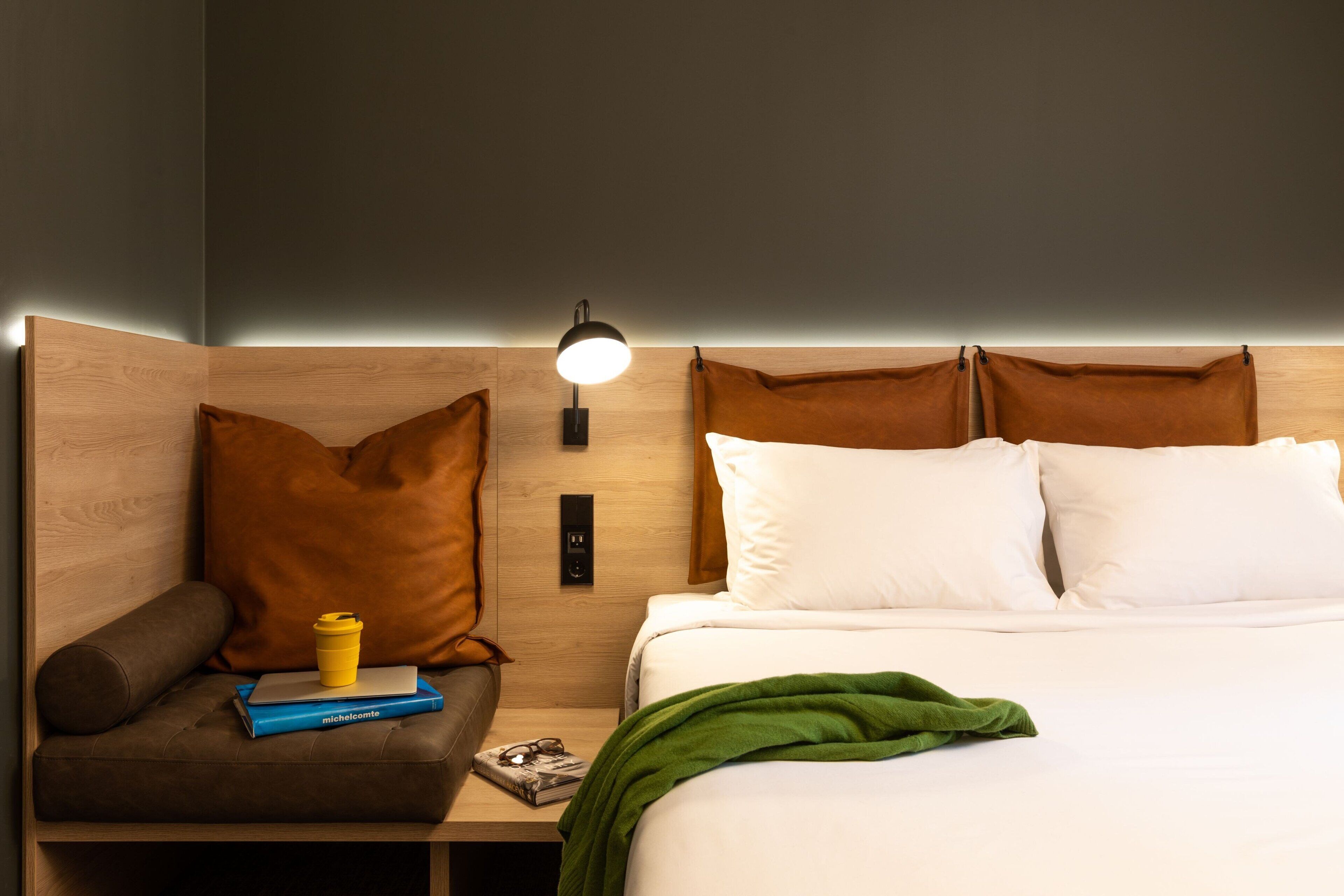 A hotel room with a wooden headboard, a bedside lamp, a brown leather chair, a green throw blanket, and a small table with a book and a cup.