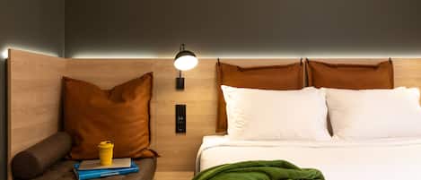 A hotel room with a wooden headboard, a bedside lamp, a brown leather chair, a green throw blanket, and a small table with a book and a cup.