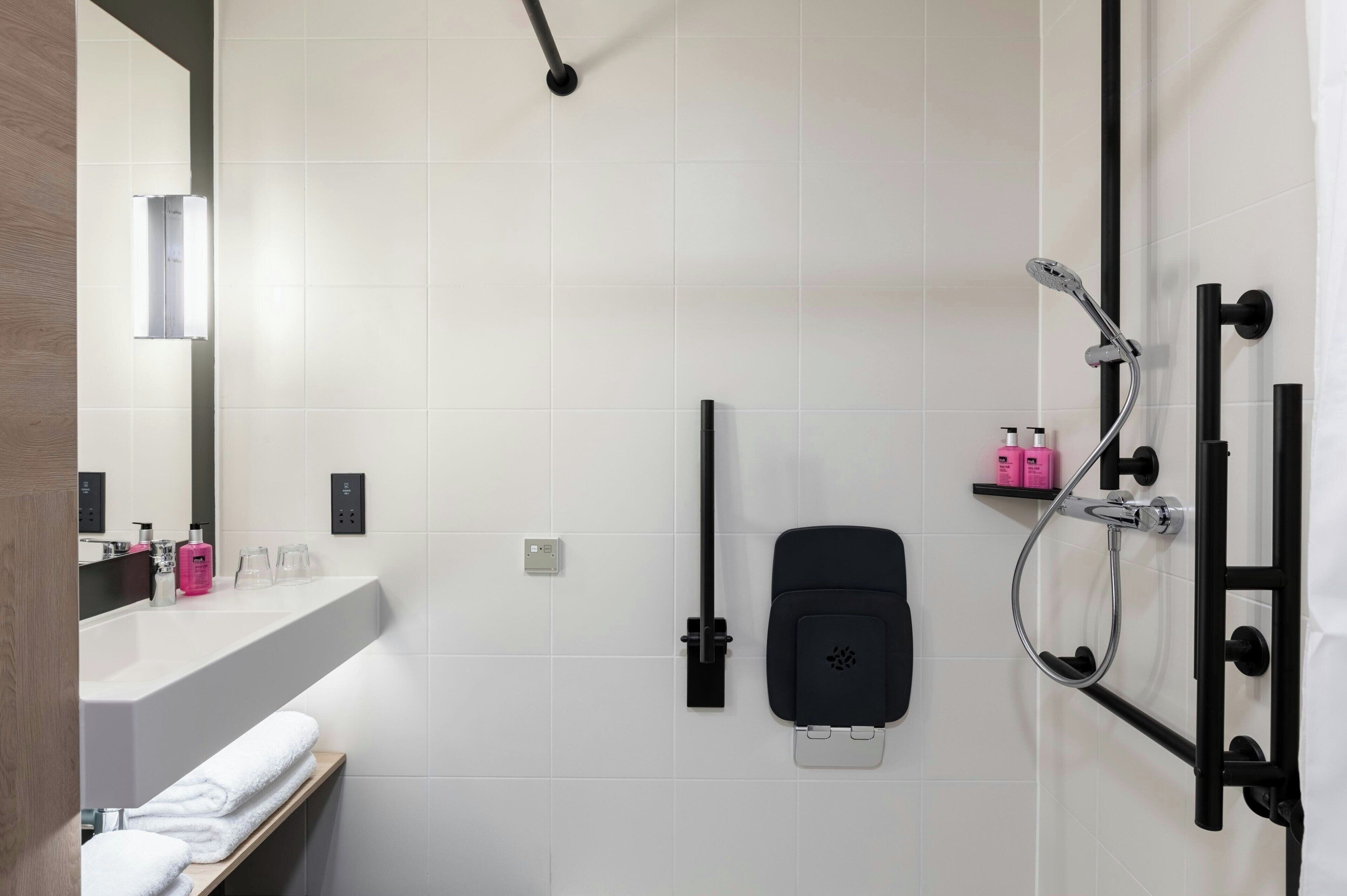 A modern bathroom with a large white sink, a black towel rack, a handheld showerhead, and a black soap dispenser.