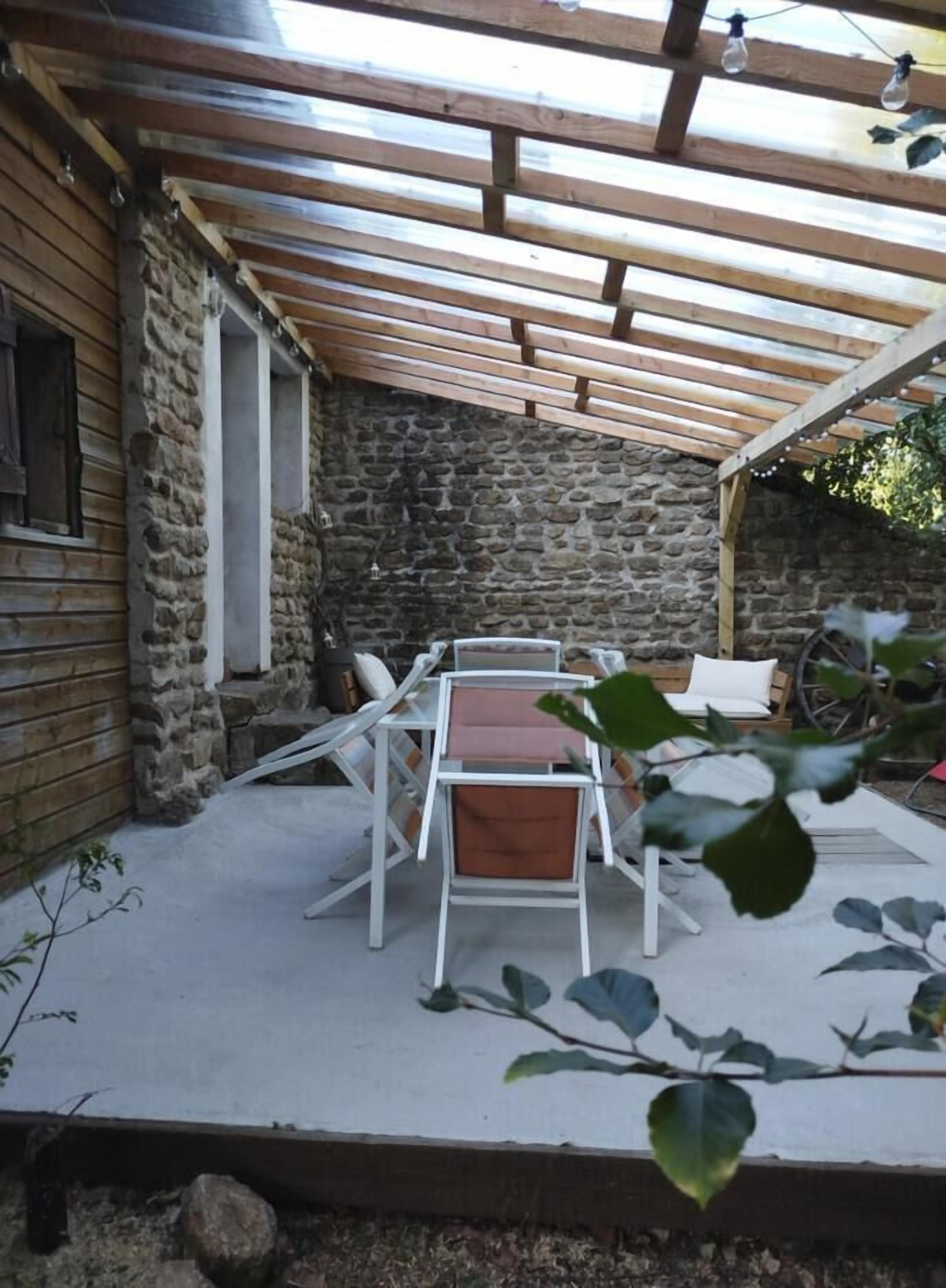 A covered outdoor patio with a stone wall, a wooden table with chairs, and a rustic aesthetic.