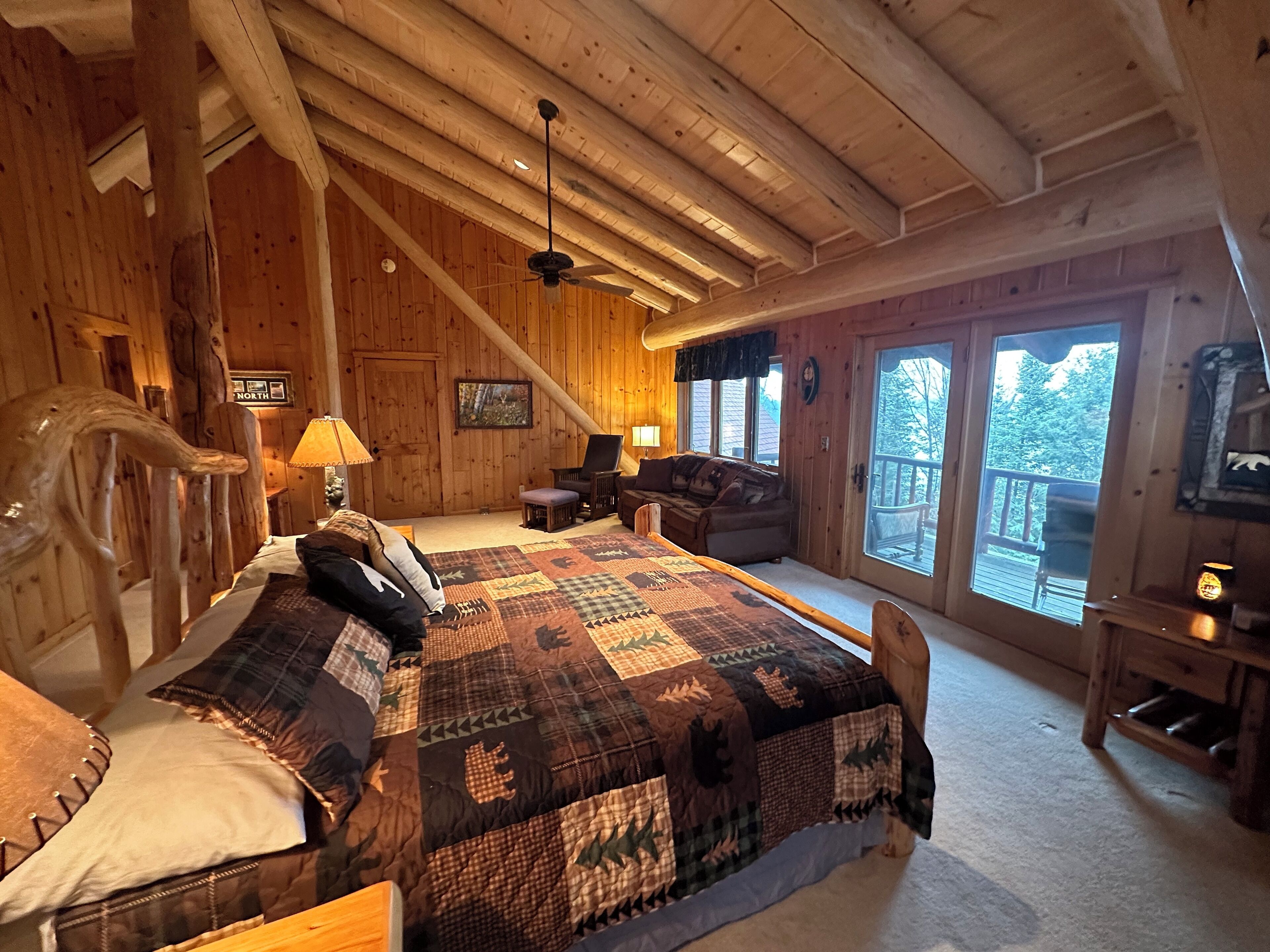 Un dormitorio tipo cabaña de madera con una cama grande, una silla, un escritorio y un balcón con vistas al exterior.