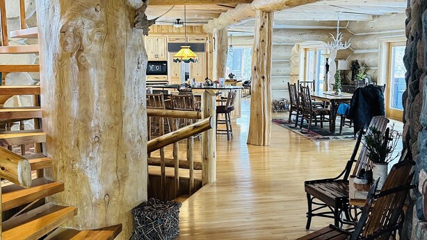 Un intérieur spacieux de chalet en bois, avec un escalier en bois, un coin repas et une cuisine dotée d’un luminaire suspendu.