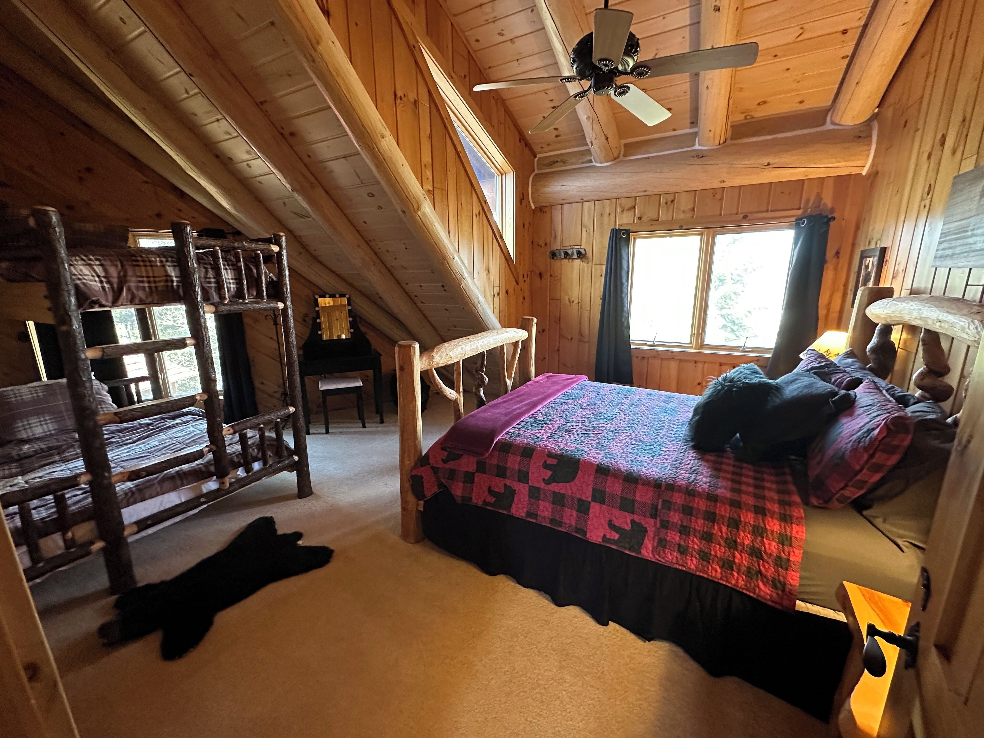 A cozy cabin room with a bunk bed, a single bed with a colorful quilt, a ceiling fan, and a window with curtains.