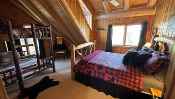 A cozy cabin room with a bunk bed, a single bed with a colorful quilt, a ceiling fan, and a window with curtains.