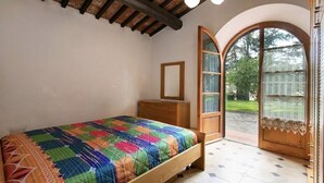 A bedroom with a wooden bed, a wooden nightstand, a mirror, and a large arched window with a view of a garden. - Maison Antoinette - Violet (Toscana)