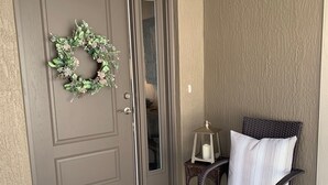 Une porte double en bois brun ornée d’une guirlande, une chaise en osier avec des coussins et une petite table sur laquelle est posée une bougie.