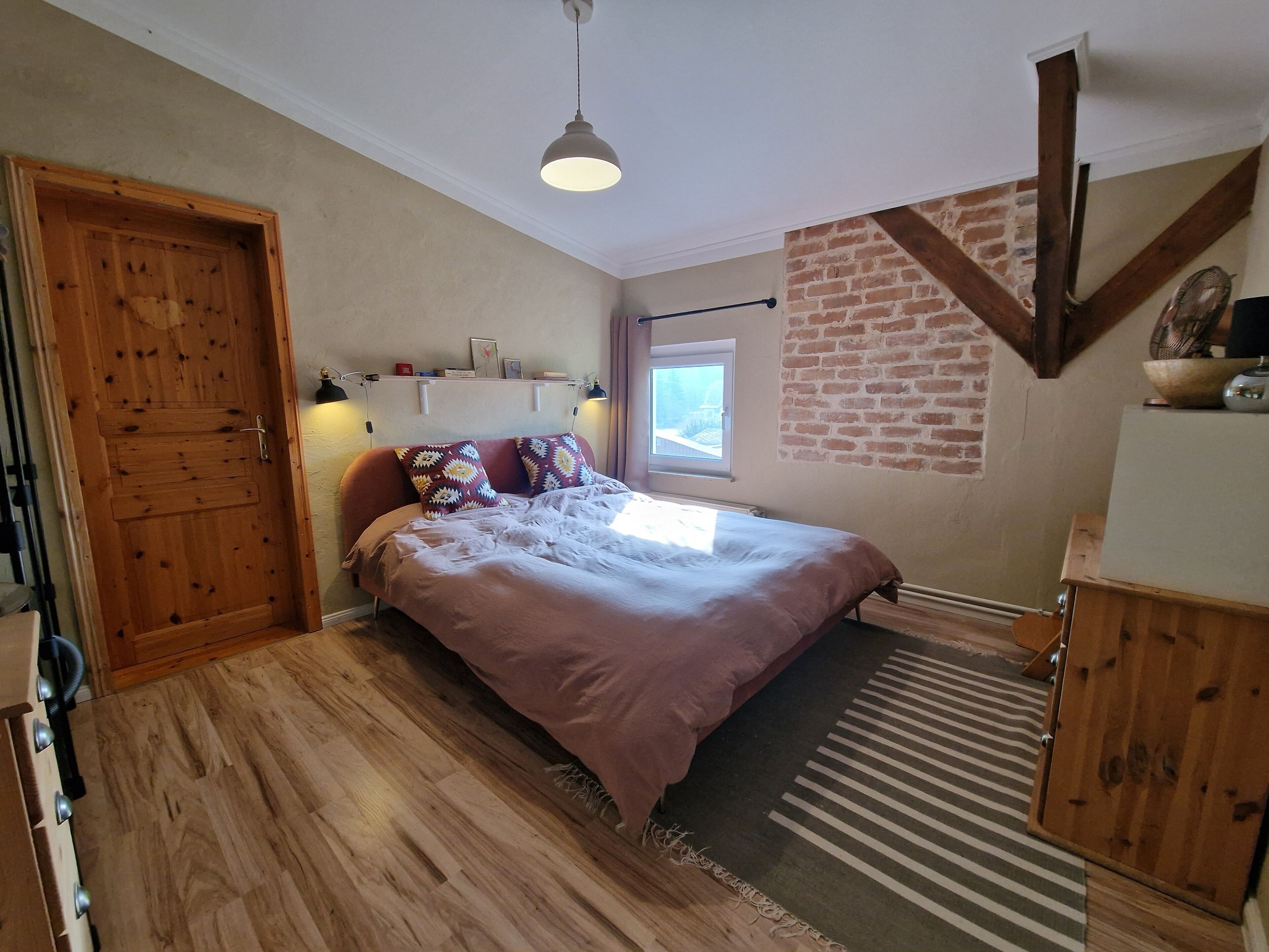 Een slaapkamer met een houten deur, een bed met gekleurde kussens, een hanglamp, een raam en een bakstenen muur.