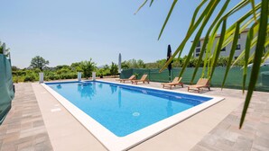Une piscine rectangulaire dont le bord est recouvert de carrelage, entourée d’une terrasse également carrelée. Quatre chaises longues en bois sont alignées le long du bord de la piscine.