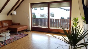 A modern living room with a brown leather sofa, a colorful rug, a large potted plant, and a sliding glass door leading to a balcony.