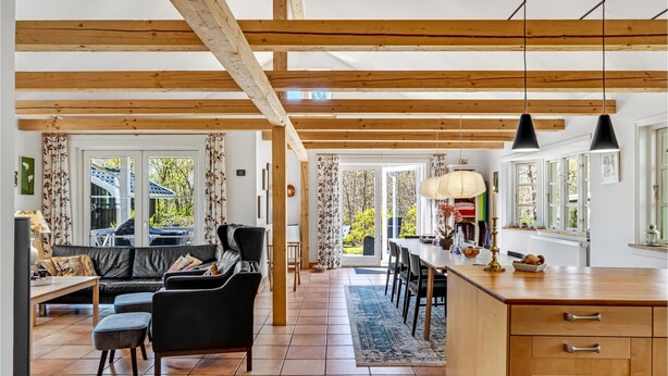 A spacious living area with a sofa, chairs, and a dining table. There is a kitchen area with wooden cabinets and a large window. - Cozy home in Græsted with sauna (Græsted)