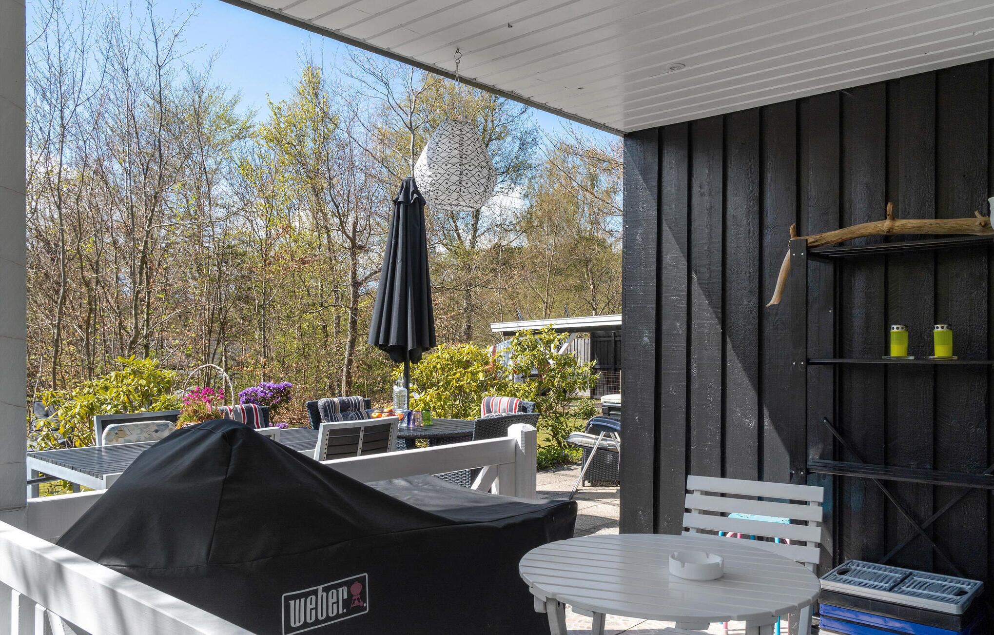 An outdoor patio area with a covered seating area, a grill, and a view of trees and a building in the background.