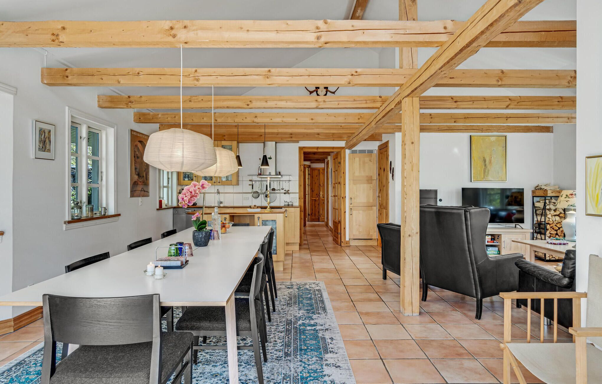 Een ruime keuken met houten balken, een eethoek met een lange tafel en stoelen, en een zithoek met een bank en televisie.