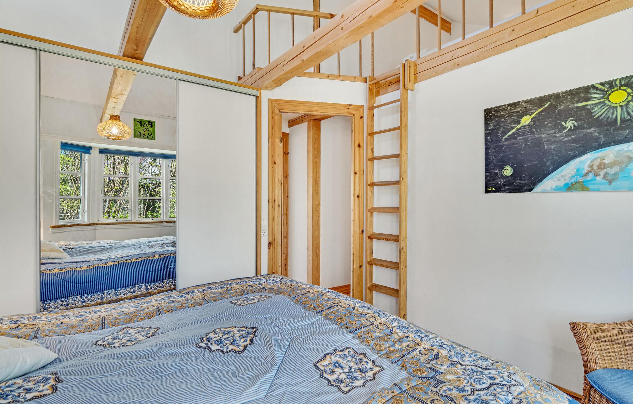 Une chambre avec un grand lit, des poutres en bois, une échelle, une fenêtre donnant sur la forêt et un tableau accroché au mur.