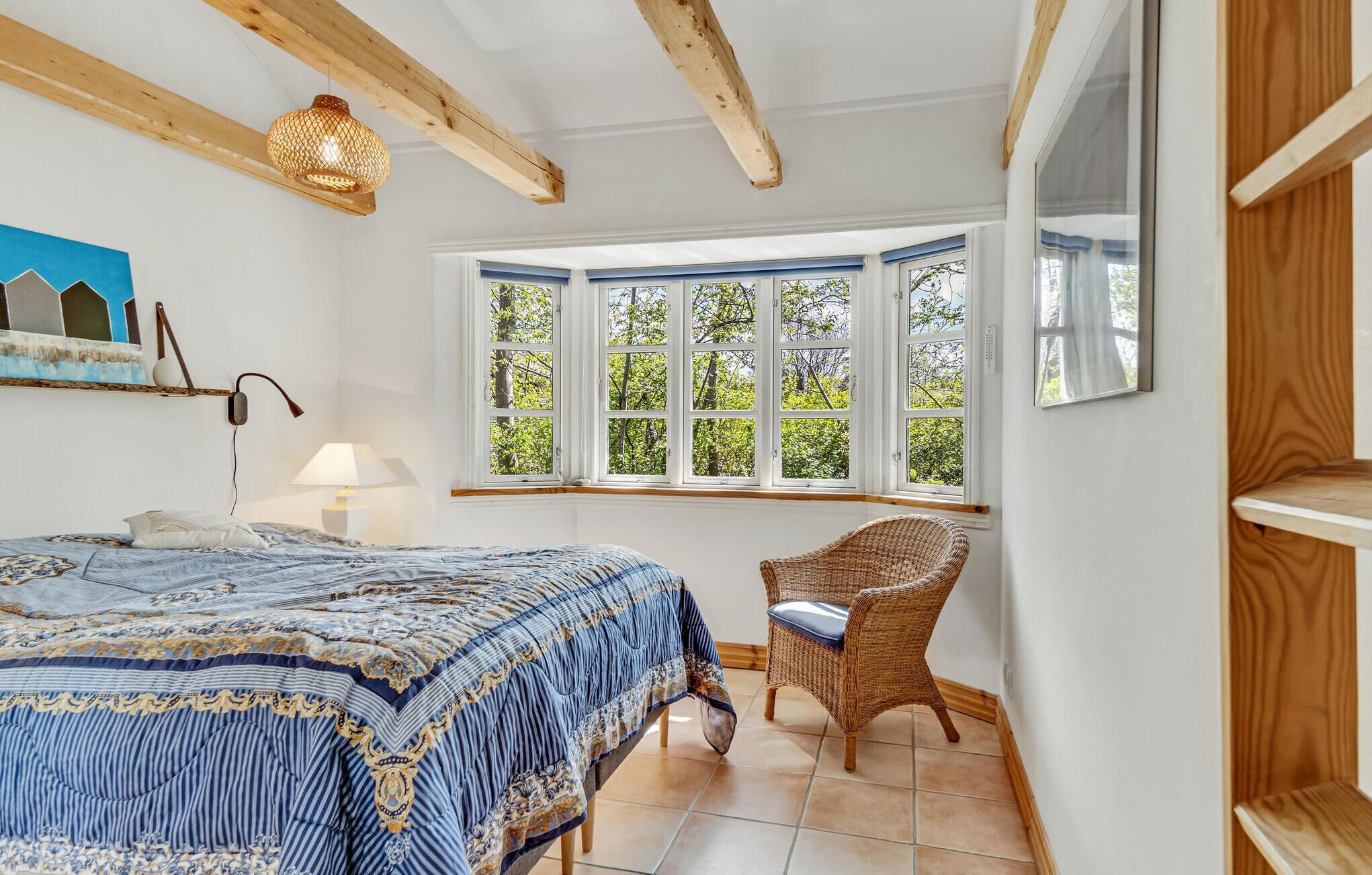 A bedroom with a large bed, a wicker chair, a window with multiple panes, and a hanging light fixture.