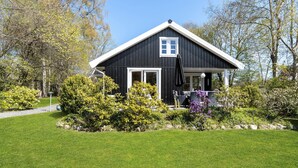 A black house with a white door and window, surrounded by a well-maintained garden with various plants and a lawn. - Cozy home in Græsted with sauna (Græsted)