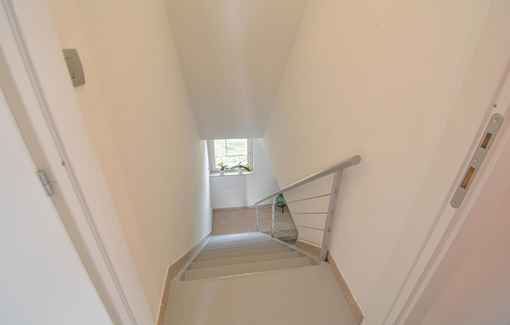 A narrow stairwell with a metal handrail, a small window at the top, and a door on the left.