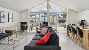 A spacious living area with a large sofa, a glass coffee table, and a dining table with chairs. There are two black armchairs, a fireplace, and a large window offering a view of the backyard.
