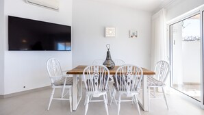 A dining area with a wooden table and white chairs, a flat-screen TV mounted on the wall, and a window with a view of the outside. - Casa do Limoeiro 54 LBC by CoolHouses (Praia da Luz)