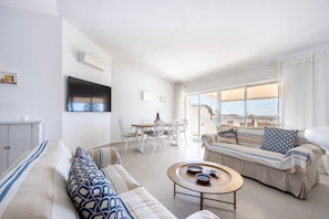 A modern living room with a sofa, a dining table with chairs, a TV mounted on the wall, and a balcony with a view.