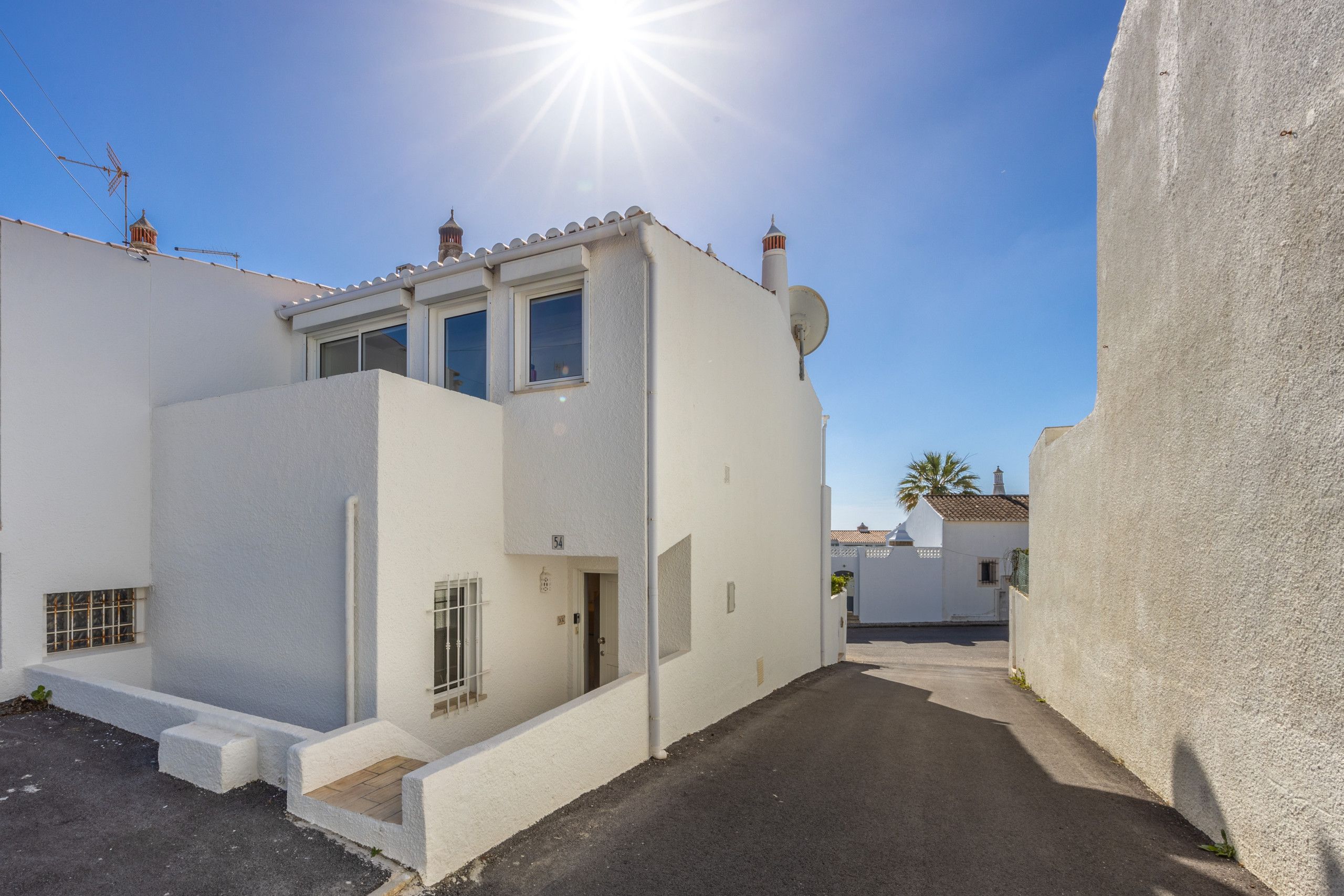 Une rue étroite bordée de bâtiments blancs, un ciel dégagé et un soleil éclatant.