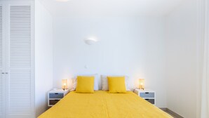 A hotel room with a bed, two bedside tables, a nightstand, a wall-mounted light, and a door with louvered panels. - Casa do Limoeiro 54 LBC by CoolHouses (Praia da Luz)