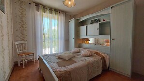 A bedroom with a bed, a chair, a wardrobe, and a window with curtains. - Maison côte sauvage. classement 3* (Quiberon)