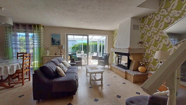 A living room with a sofa, coffee table, and a fireplace. There is a dining area with a table and chairs. The room has large windows and a staircase. - Maison côte sauvage. classement 3* (Quiberon)