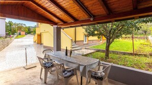 An outdoor patio with a table and chairs, a covered area with a wooden ceiling, and a view of a yellow building and greenery. - 2 bedroom nice apartment in Cernik (Cernik)