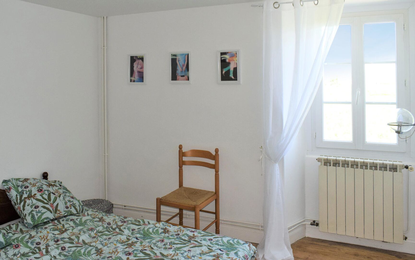 Een slaapkamer met een bed, een houten stoel, een radiator en drie foto’s in lijsten aan de muur.