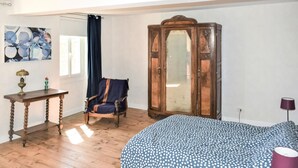 Een kamer met een houten vloer, een bed met een stippenovertrek, een stoel, een houten kast, een tafel met een lamp en een schilderij aan de muur.