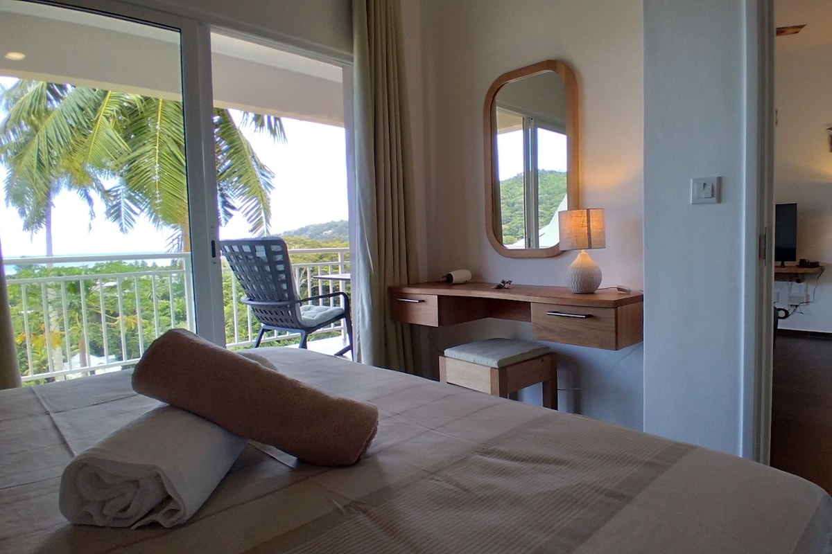 Habitación de hotel con cama, escritorio, silla, espejo y vista a palmeras.