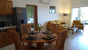 A modern kitchen with wooden cabinets, a dining area with a table set for two, and a living room with a sofa and armchairs. - La Trouvaille Holiday Chalets (Mahé Island)