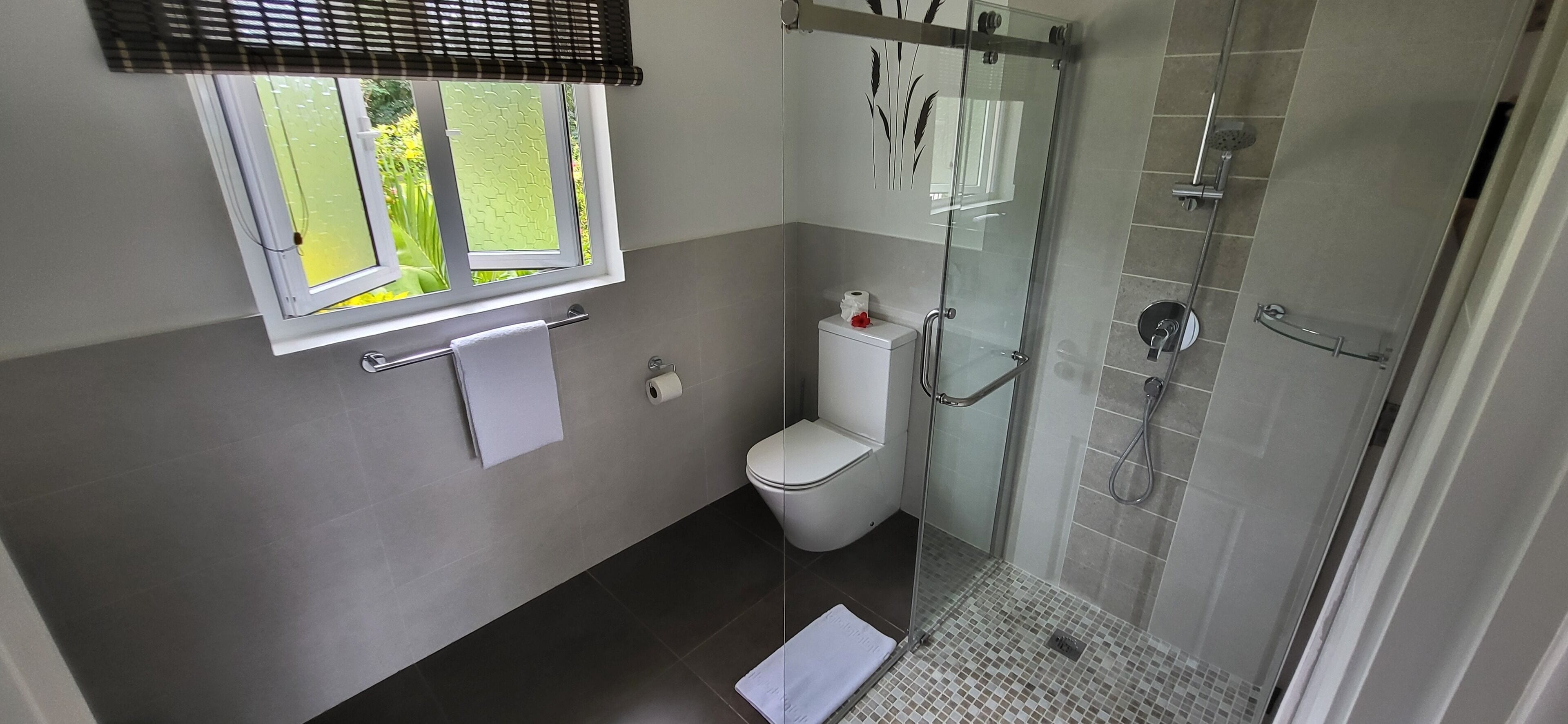 A bathroom with a toilet, a glass shower enclosure, a window with blinds, and a towel rack.