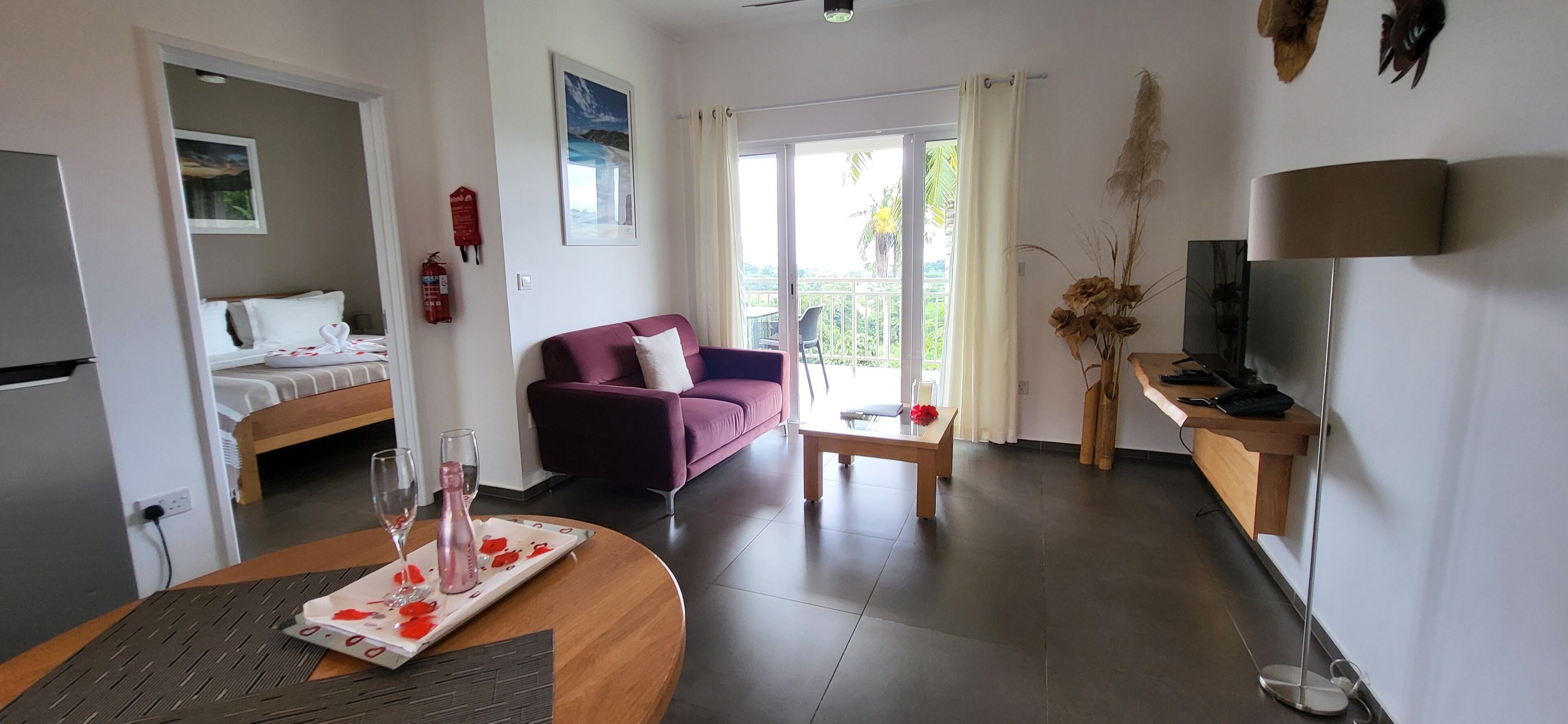 A hotel room with a bed, a purple sofa, a dining table with a placemat, a TV, and a balcony with a view.