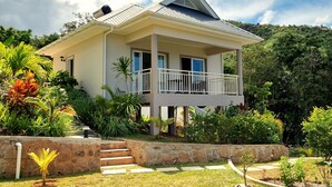 A two-story house with a balcony, surrounded by lush greenery and a stone retaining wall. - La Trouvaille Holiday Chalets (Mahé Island)