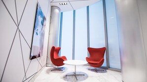 Un intérieur moderne avec deux chaises rouges disposées autour d’une petite table blanche, de grandes fenêtres et un motif géométrique sur le mur.