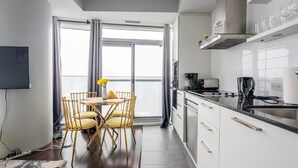 Une cuisine moderne avec des meubles blancs, un plan de travail noir et une machine à café. On y trouve un coin repas avec une table ronde et des chaises, ainsi qu’une grande fenêtre avec des rideaux.